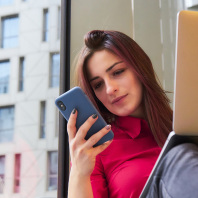 Eine Frau sitzt an einem Fenster mit einem aufgeklappten Laptop auf den Knien und einem Handy in der Hand.
