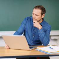 Lehrer am Pult vor Tafel blickt in ein Laptop.