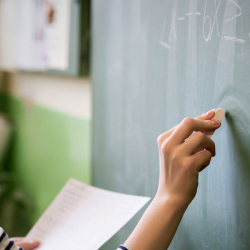 Junge Lehrkraft schreibt etwas mit Kreide an eine Tafel in einem Klassenzimmer. © Andrea Obzerova / AdobeStock