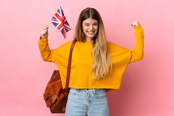 Eine junge Person hält eine britische Flagge in die Höhe, mit der anderen Hand formt sie eine Faust. Sie hat langes blondes Haar, trägt einen gelben Pullover und eine Jeans. Der Hintergrund ist rosa.