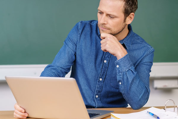 Lehrer am Pult vor Tafel blickt in ein Laptop.