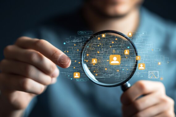 Eine Person hält eine Lupe, die sich auf digitale Symbole in einem modernen Arbeitsumfeld fokussiert und damit Analyse sowie Einblicke in Trends der Netzwerk- und Kommunikationstechnologie symbolisiert.