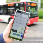 Eine Hand hält ein Smartphone mit der geöffneten Nürnberg Mobil-App, im Hintergrund steht ein Bus.