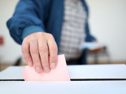 Foto von einem Mann mit einem Wahl·schein an einer Wahl·urne.