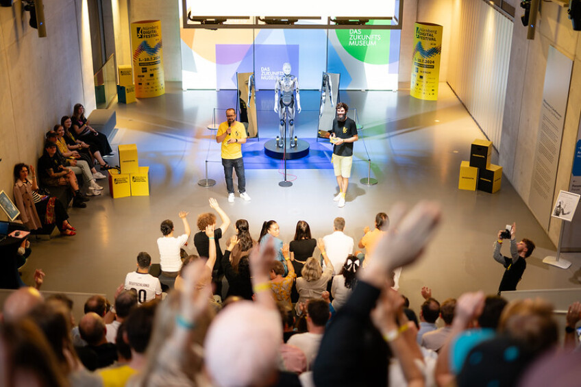 Bild © endlosmedia Abschlussveranstaltung des Nürnberg Digital Festivals 2024. Ein Roboter und zwei Personen stehen auf einer Bühne im Hintergrund. Im Vordergrund sitzen Teilnehmer der Veranstaltung., Bild © endlosmedia