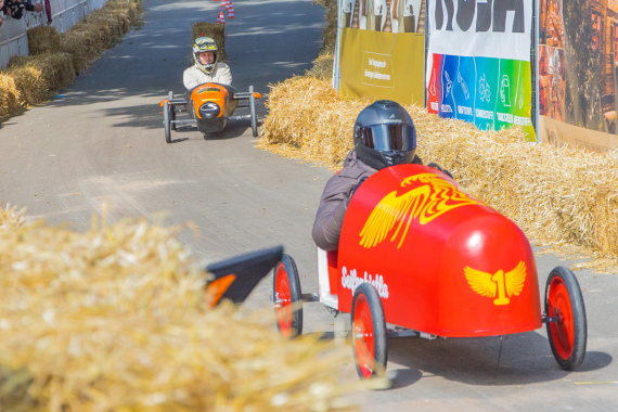 Zwei Seifenkisten beim Rennen im Burggraben. Die Strecke ist gesäumt von Strohballen. Besucher am Fahrbahnrand feuern die Fahrer an.