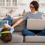 Eine Mutter mit Laptop sitzt neben einem Kind mit Tablet auf dem Sofa.