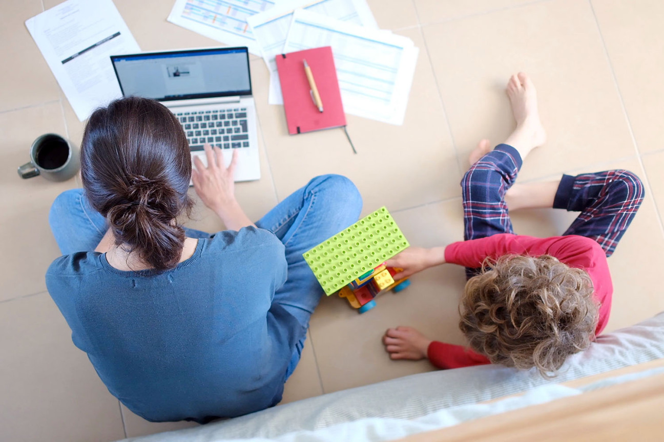 Eine Frau sitzt mit einem Kind auf dem Fußboden. Sie tippt auf einem Laptop, drumherum liegen Notizen. Daneben spielt ein kleiner Junge mit einem Leogauto., Bild © DawDunia / AdobeStock