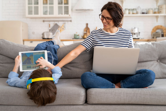 Eine Mutter mit Laptop sitzt neben einem Kind mit Tablet auf dem Sofa.