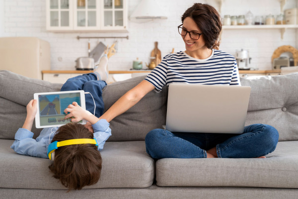 Foto: Eine Mutter mit Laptop sitzt neben einem Kind mit Tablet auf dem Sofa.