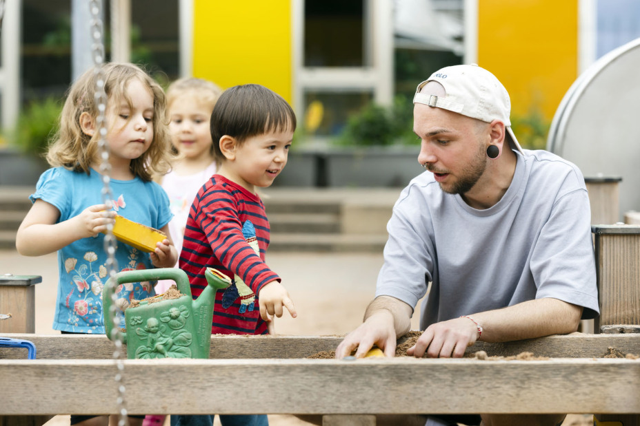 Ein Erzieher aus einer Nürnberger Kita spielt mit Kindern im Sandkasten.