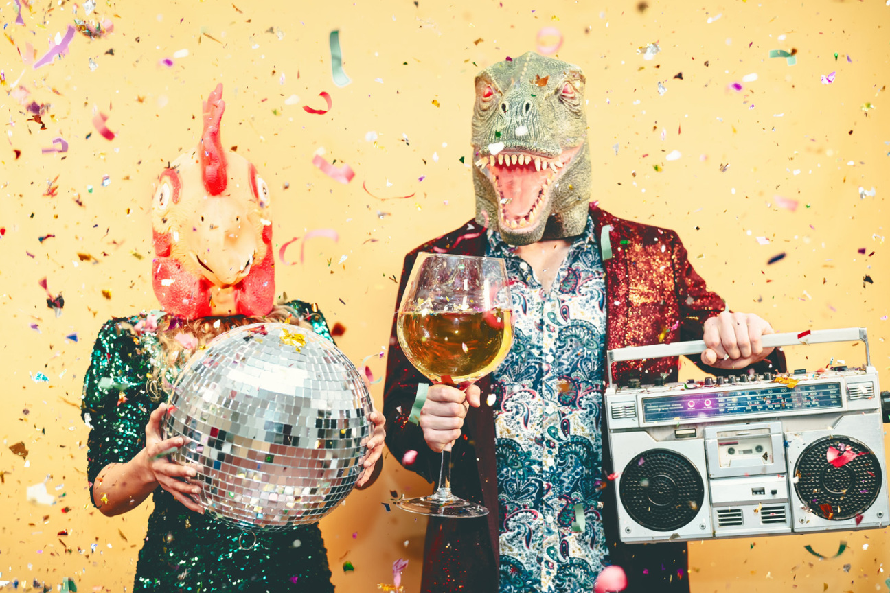 Ein Mann und eine Frau, verkleidet mit einer Dino-Maske und einer Hahn-Maske halten eine Diskokugel und einen Ghettoblaster. Lametta rieselt von der Decke., Bild © Alessandro Biascioli / Adobestock/320291724