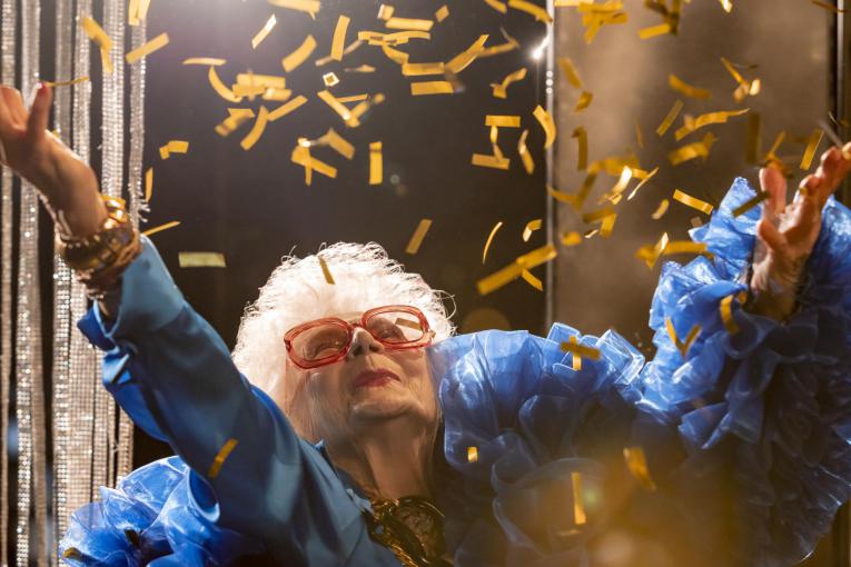 Eine ganz in blau gekleidete ältere Person mit weißen Haaren und roter Brille wirft goldene Konfettistreifen in die Luft.