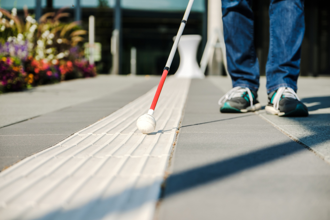 Bild © Andi Weiland / www.gesellschaftsbilder.de Blindenstock tastet über Leitstreifen, dahinter Sneakers und Jeans der blinden Person, Bild © Andi Weiland / www.gesellschaftsbilder.de