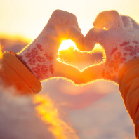 Eine Frau mit Handschuhen formt mit ihren Händen ein Herz, durch das die Wintersonne hindurch scheint.