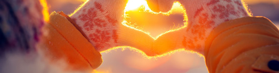 Eine Frau mit Handschuhen formt mit ihren Händen ein Herz, durch das die Wintersonne hindurch scheint.