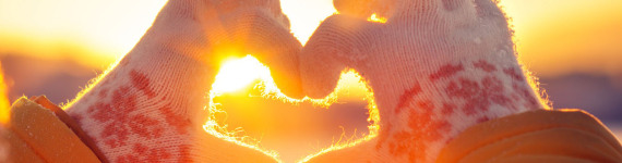 Eine Frau mit Handschuhen formt mit ihren Händen ein Herz, durch das die Wintersonne hindurch scheint.