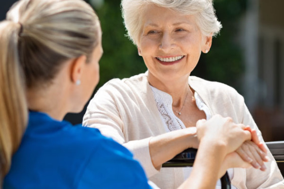 Foto von 2 Frauen: eine junge Frau und eine alte Frau. Die junge Frau ist eine Pflegerin. Die alte Frau lächelt.