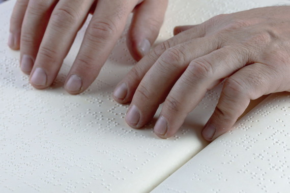 Zwei männliche Hände in Großaufnahme, die über ein Buch mit Brailleschrift gleiten.