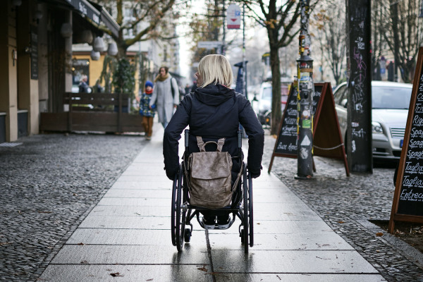 Eine Frau sitzt in einem Rollstuhl und fährt einen Gehweg entlang.