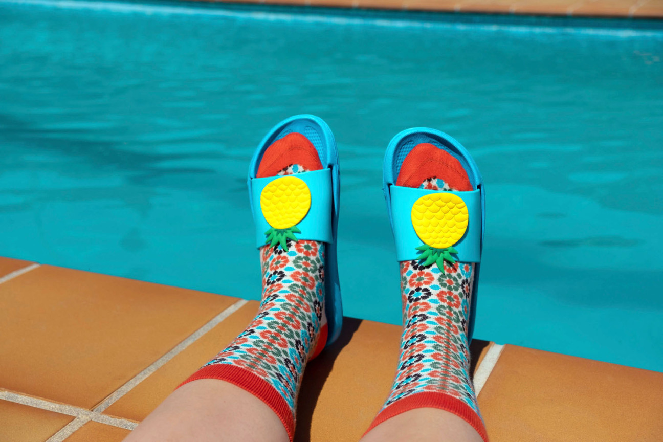 Füße mit bunten Socken und blauen Badesandalen am Rand eines Schwimmbades., Bild © Ana / Adobe Stock