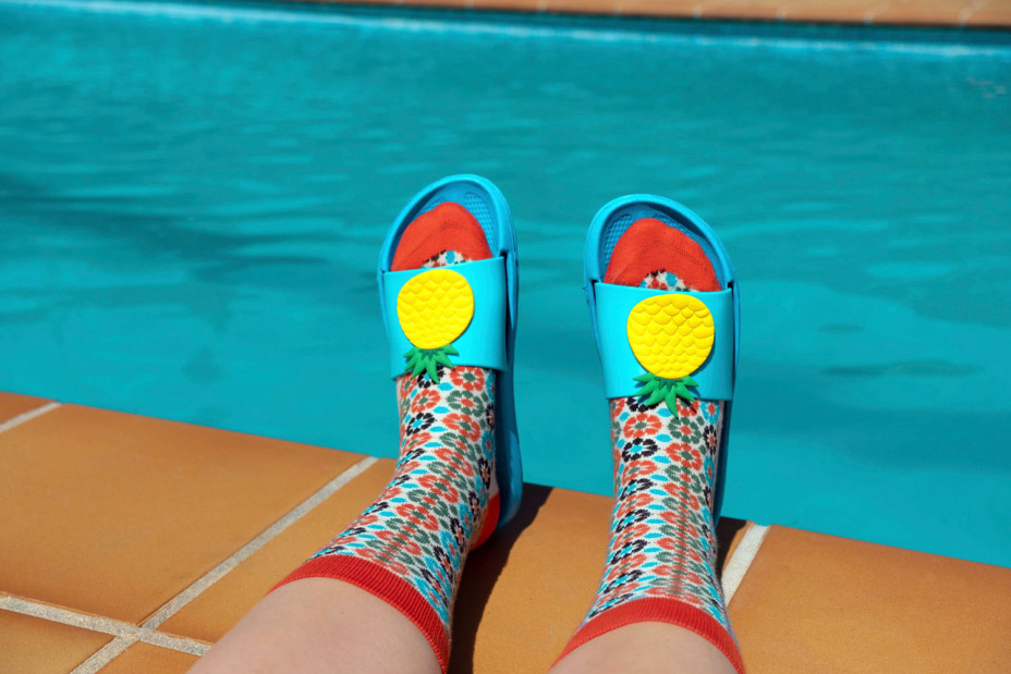 Füße mit bunten Socken und blauen Badesandalen am Rand eines Schwimmbades.