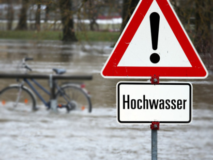 Foto von einem Warn·schild Hoch·wasser vor einem überfluteten Gebiet.