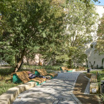 Blick auf den Pocket Park Peststadel in Nürnberg. Durch die Mitte des Parks führt ein Weg. Er ist links und rechts von Bäumen, Büschen und grün gesäumt. Entlang des Weges stehen bequeme Liegebänke aus Holz. Sie laden zum Ausruhen ein.