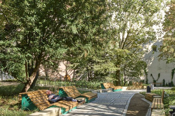 Blick auf den Pocket Park Peststadel in Nürnberg. Durch die Mitte des Parks führt ein Weg. Er ist links und rechts von Bäumen, Büschen und grün gesäumt. Entlang des Weges stehen bequeme Liegebänke aus Holz. Sie laden zum Ausruhen ein.