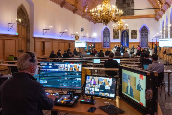 Foto von einem Digitalen Bürger·dialog. Vorn im Bild steht ein Mann vor 3 Bildschirmen. Hinten im Bild sieht man einzelne Personen.