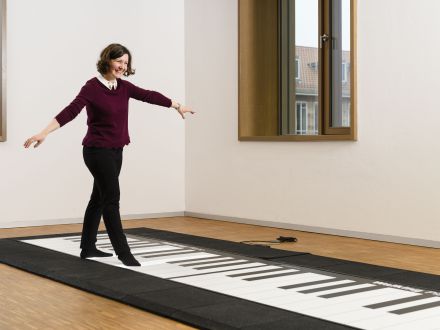 Das Bild zeigt eine Frau auf dem Boden·klavier. Das Klavier ist groß und flach. Und das Klavier ist direkt am Boden. Um das Boden∙klavier ist eine Absperrung aufgestellt.