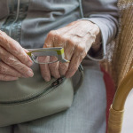 Die Hände einer älteren Dame halten eine Handtasche und eine Brille.