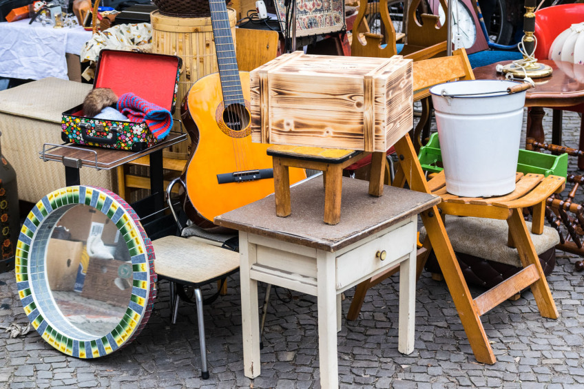 Flohmarktgegenstände bestehend aus kleinen Tischen, Hocker, Klappstuhl, Eimer, Spiegel, Gitarre, und Lampenschirm sind zu einem kunterbunten Stapel aufgetürmt., Bild © AdobeStock