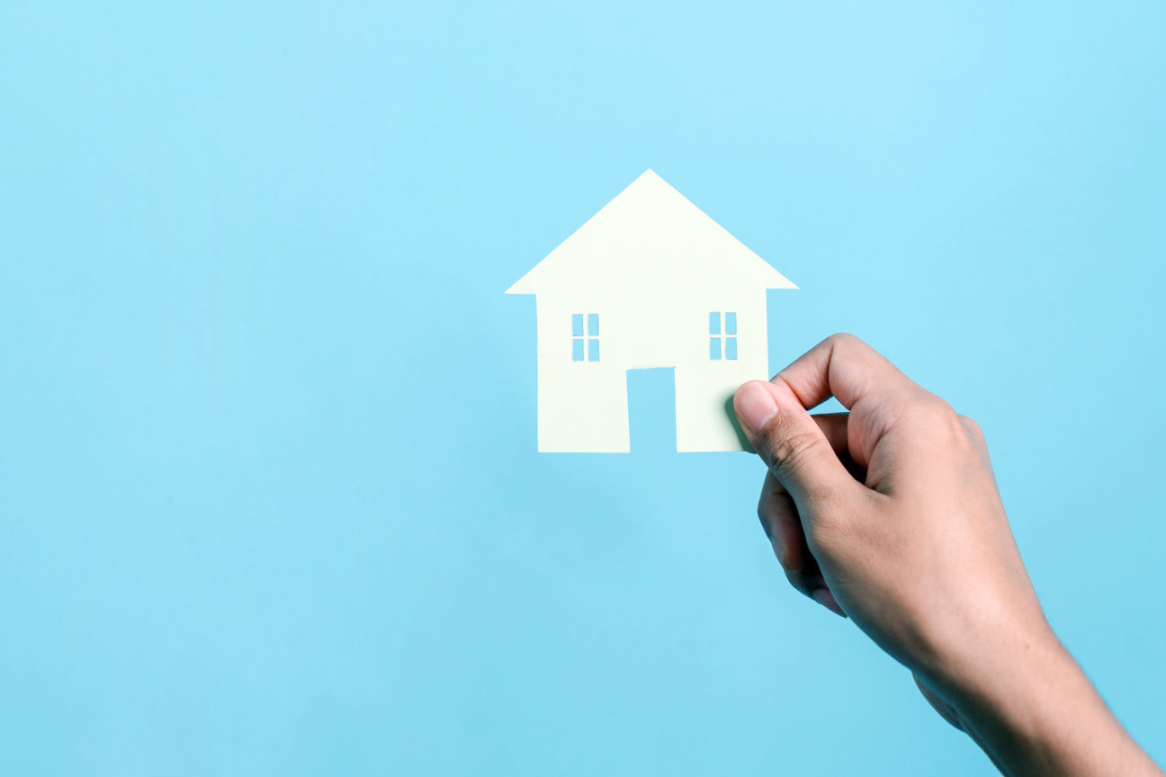 Symbolbild: Eine Hand hält ein aus einem Stück Papier ausgeschnittenes Haus vor blauem Hintergrund., Bild © Kiattisak / AdobeStock