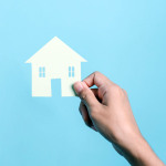 Symbolbild: Eine Hand hält ein aus einem Stück Papier ausgeschnittenes Haus vor blauem Hintergrund.