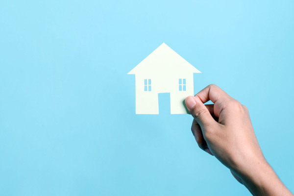 Symbolbild: Eine Hand hält ein aus einem Stück Papier ausgeschnittenes Haus vor blauem Hintergrund.