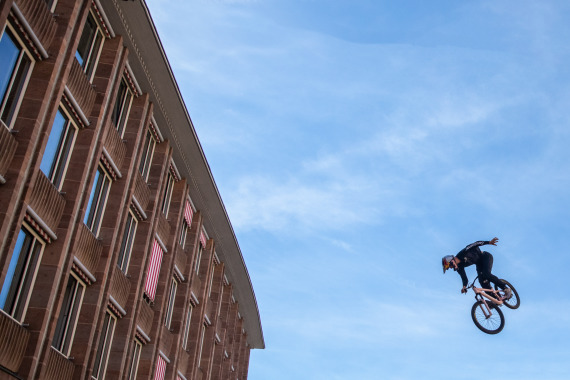 Mountainbiker beim Sprung in der Luft, links daneben das Rathaus, blauer Himmel.