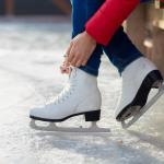 Eine Person sitzt auf einer Bank am Rande einer Eisfläche und bindet Damenschlittschuhe.
