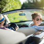 Junges Paar fährt an einem sonnigen Sommertag mit einem Cabrio auf einer Landstraße.