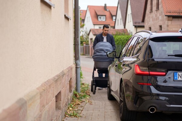 Ein Mann schiebt einen Kinderwagen und versucht, an einem auf dem Gehweg parkenden Auto vorbeizukommen.