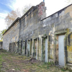 Fassade des Bahnhofs Märzfeld. In die Sandsteinmauer sind zehn Türen eingelassen. Die Mauer ist am vorderen Ende im oberen Bereich abgebrochen und nicht mehr vollständig. Die Mauer ist modrig. Im Vordergrund und Hintergrund ist sie von Grün umgeben.