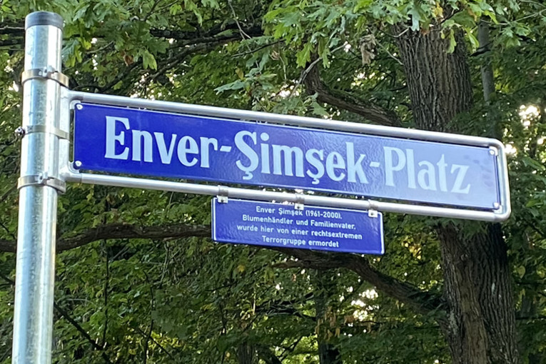 Nahaufnahme des Straßenschildes Enver-Şimşek-Platz. Es ist blau mit weißer schrift. Unterhalb des Namens befindet sich ein kleineres Schild mit der Zusatzinformatione, dass Enver Şimşek Blumenhändler und Vater war und im Jahr 2000 von einer rechtsextremen Terrorgruppe ermordet wurde.