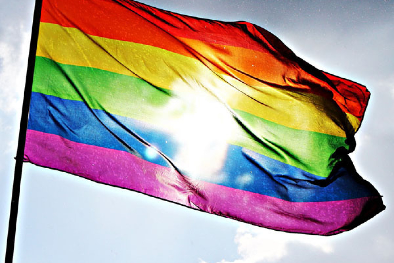 Eine Flagge weht im Wind. Sie hat die Farben eines Regenbogens bestehend aus Streifen in den Farben rot, gelb, grün, blau und violett. Dahinter ist blauer Himmel zu sehen., Bild © AdobeStock