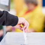 Eine Hand hält einen Wahlzettel und steckt ihr in den Schlitz einer Wahlurne.