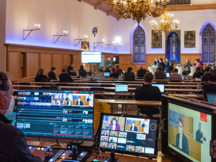 Foto von einem Digitalen Bürger·dialog. Vorn im Bild steht ein Mann vor 3 Bildschirmen. Hinten im Bild sieht man einzelne Personen.