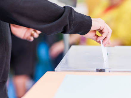 Das Bild zeigt einen Behälter für die Stimm·zettel bei der Wahl. Eine Person steckt gerade einen Stimm·zettel in den Behälter.