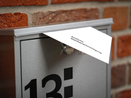 Das Bild zeigt einen Brief∙kasten an einer Haus∙wand. Aus dem Schlitz vom Brief∙kasten schaut ein Brief heraus. Der Brief ist die Wahl∙benachrichtigung vom Wahl∙amt.