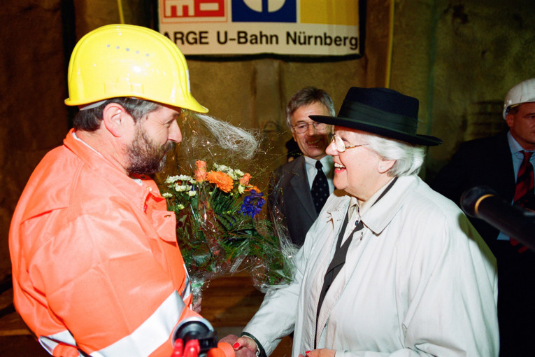Helene Jungkunz bei Feierlichkeiten anlässlich des Tunneldurchstoßes für die Linie U3 am 2. September 2001. Sie trägt einen beigen Mantel, einen schwarzen Hut und schüttelt einer Person die Hand. Die Person trägt einen gelben Bauhelm und eine orangene Schutzjacke.