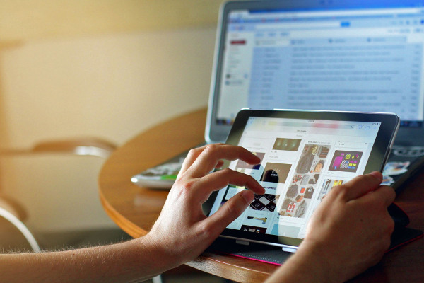 Foto: 2 Hände halten ein Tablet. Im Hintergrund ist ein Laptop.