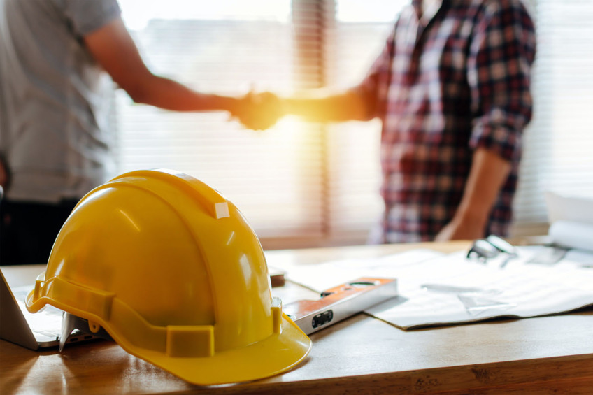 Bild © AdobeStock Zwei Männer schütteln sich die Hände. Im Vordergrund liegt ein gelber Bauhelm., Bild © AdobeStock